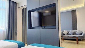 Una moderna camera d'albergo con due letti, una TV a schermo piatto fissata al muro, un divano grigio e un tavolino in legno.