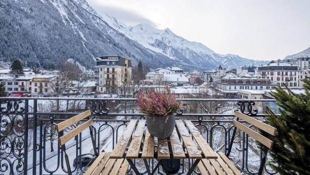 A balcony with a wooden table and chairs, a potted plant, and a view of a snow-covered mountain and town. - Nice Apt With Balcony In Chamomix (Chamonix-Mont-Blanc)
