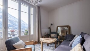A living room with a sofa, armchair, and round coffee table. Large windows offer a view of mountains. - Nice Apt With Balcony In Chamomix (Chamonix-Mont-Blanc)