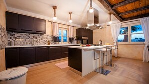 A modern kitchen with dark cabinetry, a central island, and a view of the outdoors through large windows. - Charming Villa Tomani with pool and jacuzzi (Krmed)