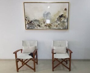 Deux chaises en bois avec des coussins blancs, placées devant un mur uni, et une grande peinture abstraite accrochée au-dessus.