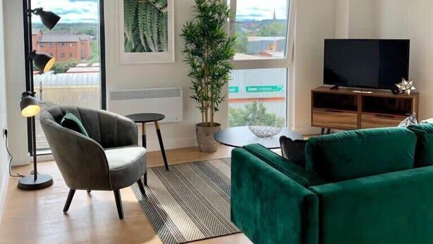 Un salon moderne avec un canapé vert, un fauteuil noir, un téléviseur à écran plat et une grande fenêtre donnant sur la ville.