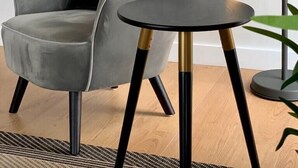 Un fauteuil gris avec un coussin vert, une petite table ronde aux pieds noirs et dorés, et un lampadaire à lumière jaune.