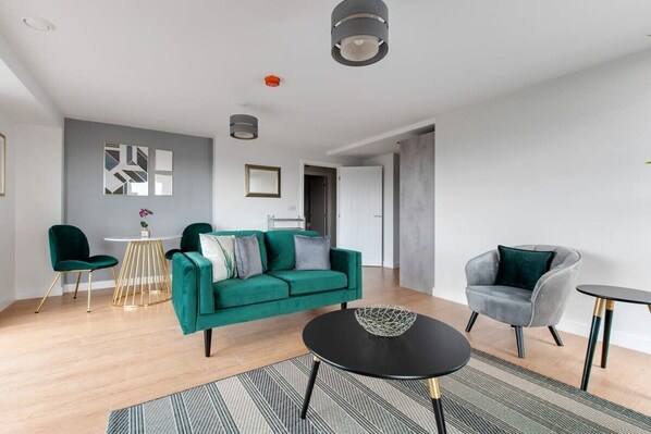 Un salon moderne avec un canapé vert, une table basse noire et un fauteuil gris. On y trouve une petite table ronde ornée d’une plante et un tableau encadré accroché au mur.