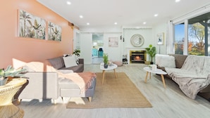 A modern living room with a sofa, armchairs, a coffee table, and a fireplace. There are two paintings on the wall, a dining area in the background, and a large window with a view of trees.