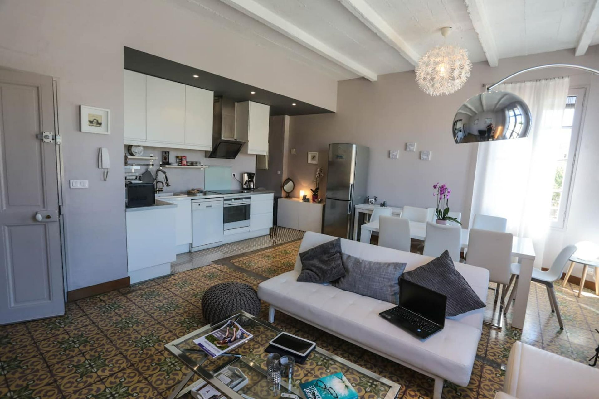 A modern living room with a sofa, coffee table, and a laptop. There is a kitchen area with white cabinets and a refrigerator.