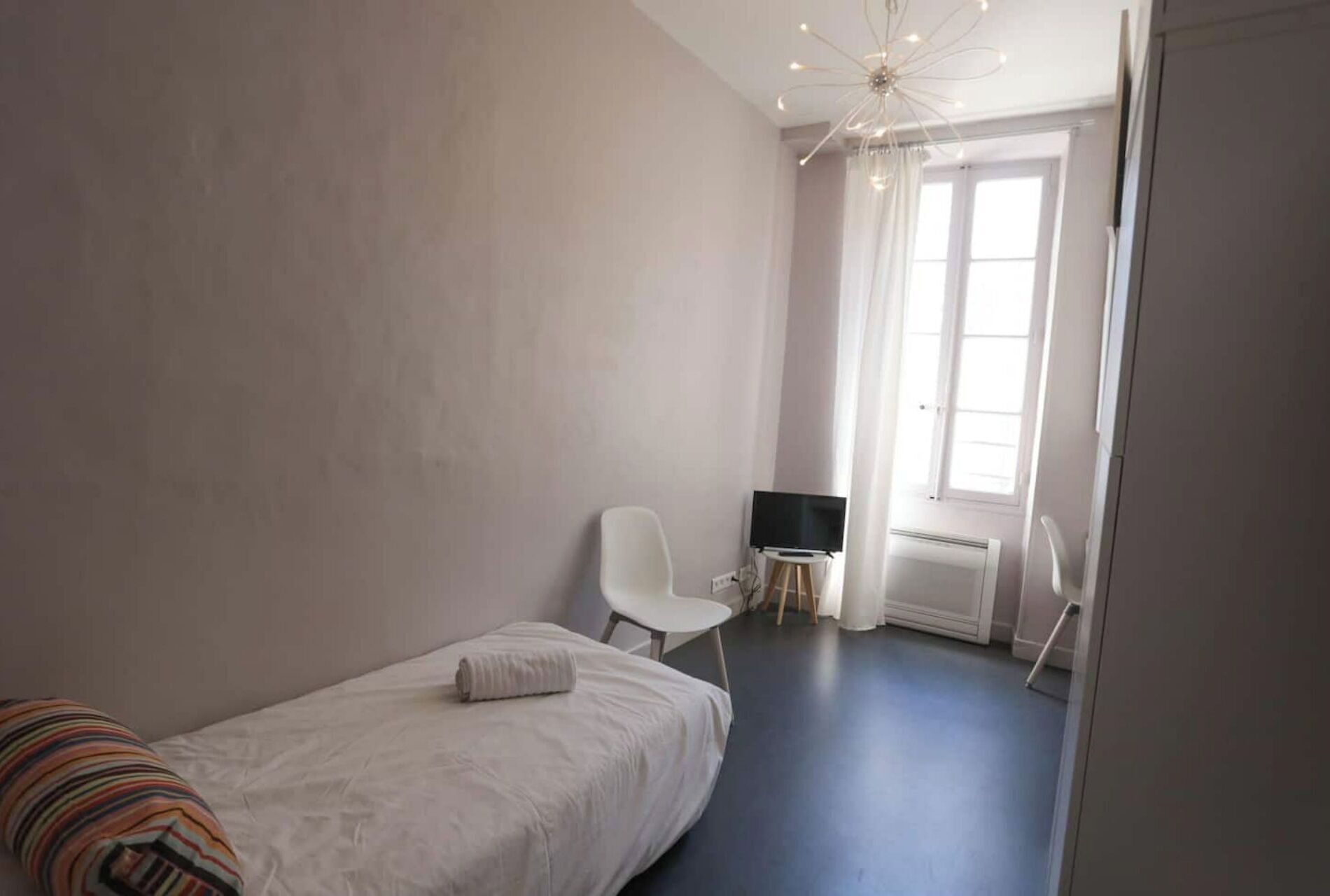 Un dormitorio con una cama individual, una silla, un televisor sobre una mesita y una ventana con cortinas.