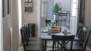 Un coin repas avec une table dressée pour quatre personnes, des chaises, une poubelle et une vue sur l’extérieur par une porte ouverte.