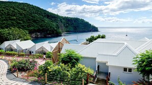 A coastal resort with white buildings, a paved walkway, and a view of the sea and a sailboat. - Les Galets Rouges Lodges & Spa (Bouillante)