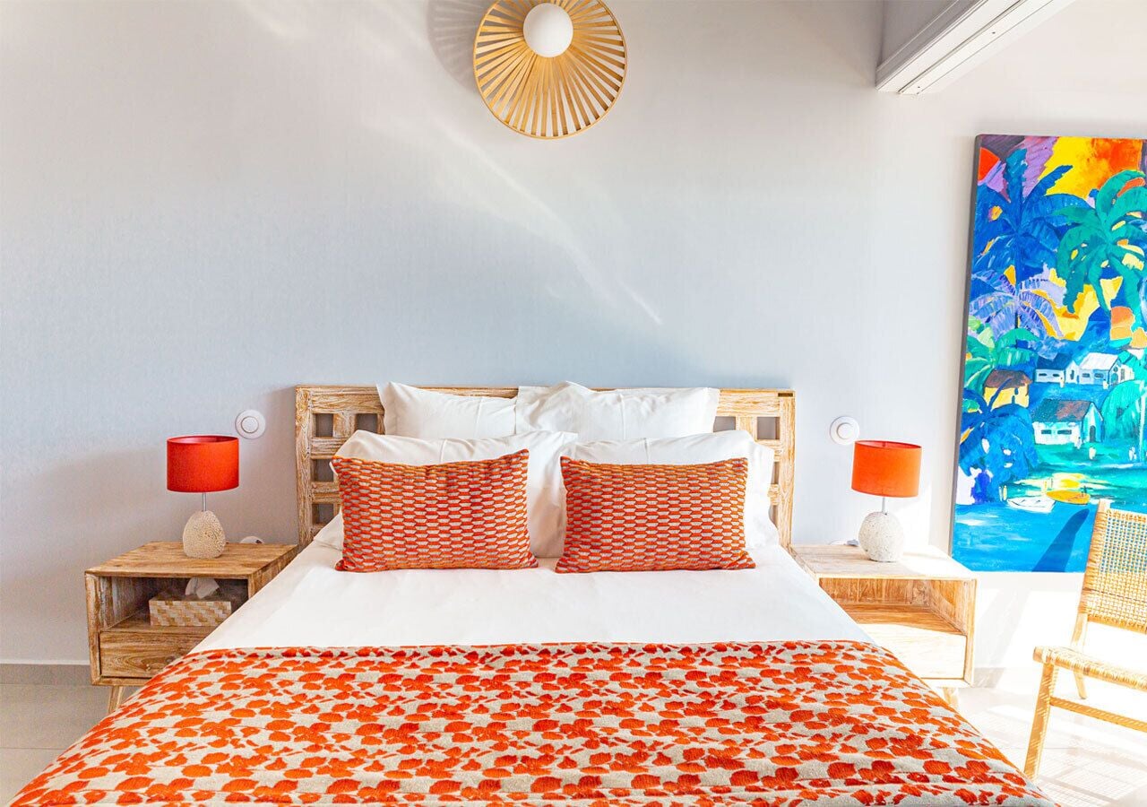 A bed with white bedding and orange patterned pillows, a wooden headboard, two nightstands with red lamps, and a painting of a tropical scene on the wall.