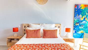 A bed with white bedding and orange patterned pillows, a wooden headboard, two nightstands with red lamps, and a painting of a tropical scene on the wall.