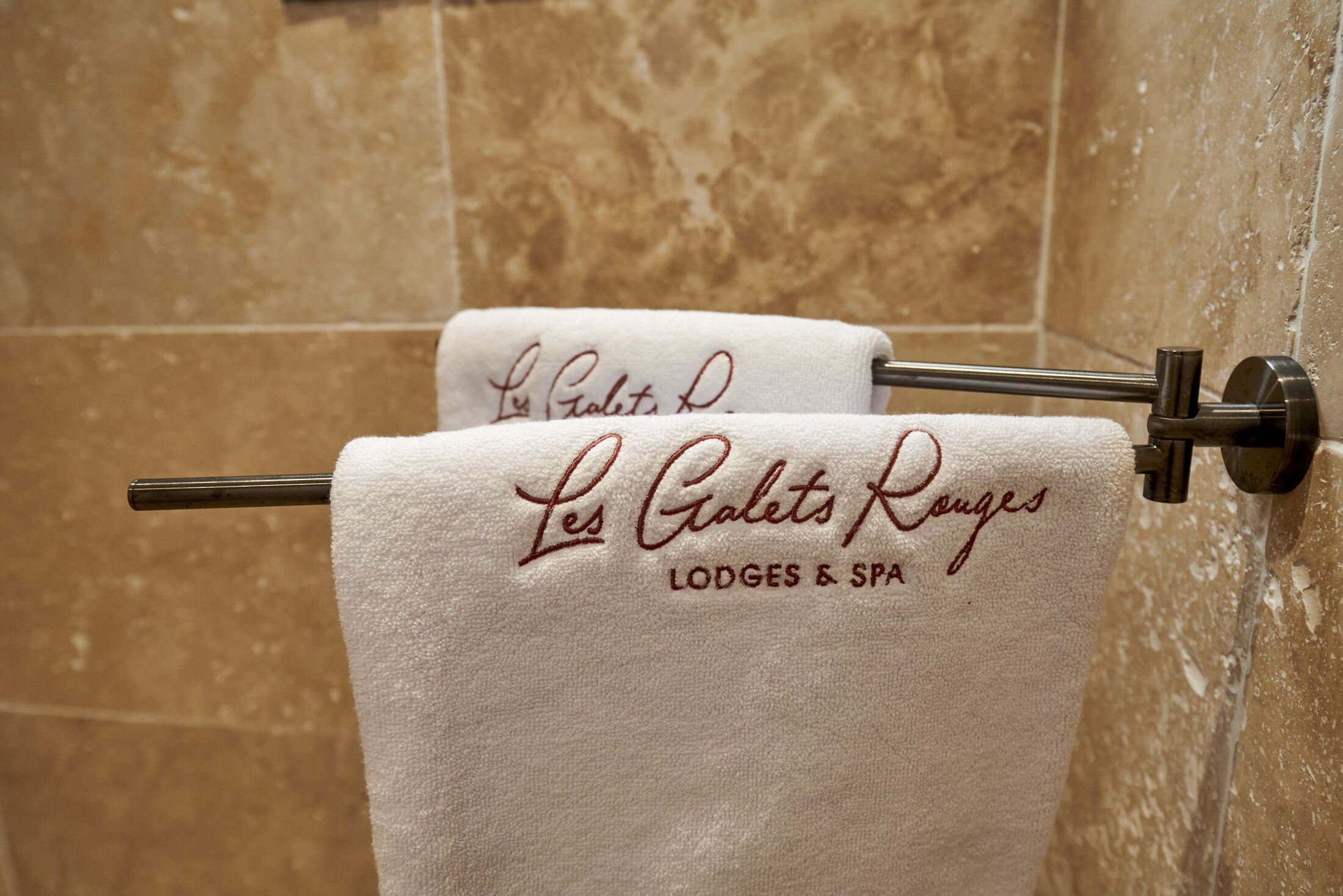 Une serviette ornée d’une broderie « Les Galets Ranges LODGES & SPA », suspendue à un porte-serviettes en métal dans une salle de bain aux carreaux beiges.