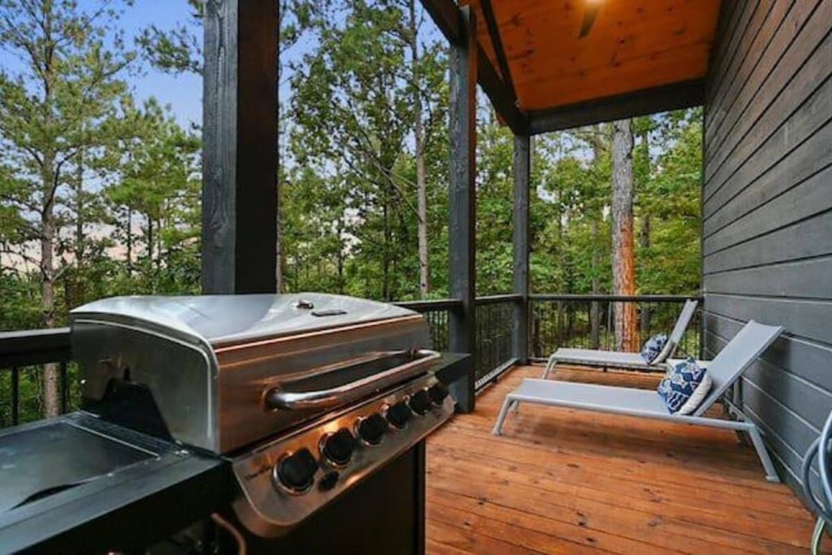 An outdoor deck with a grill, lounge chairs, and a view of trees.