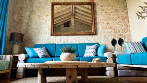 Une pièce chaleureuse avec un mur en pierre, un plafond en bois, un canapé bleu agrémenté de coussins à motifs, une table basse en bois et un miroir qui reflète le plafond.