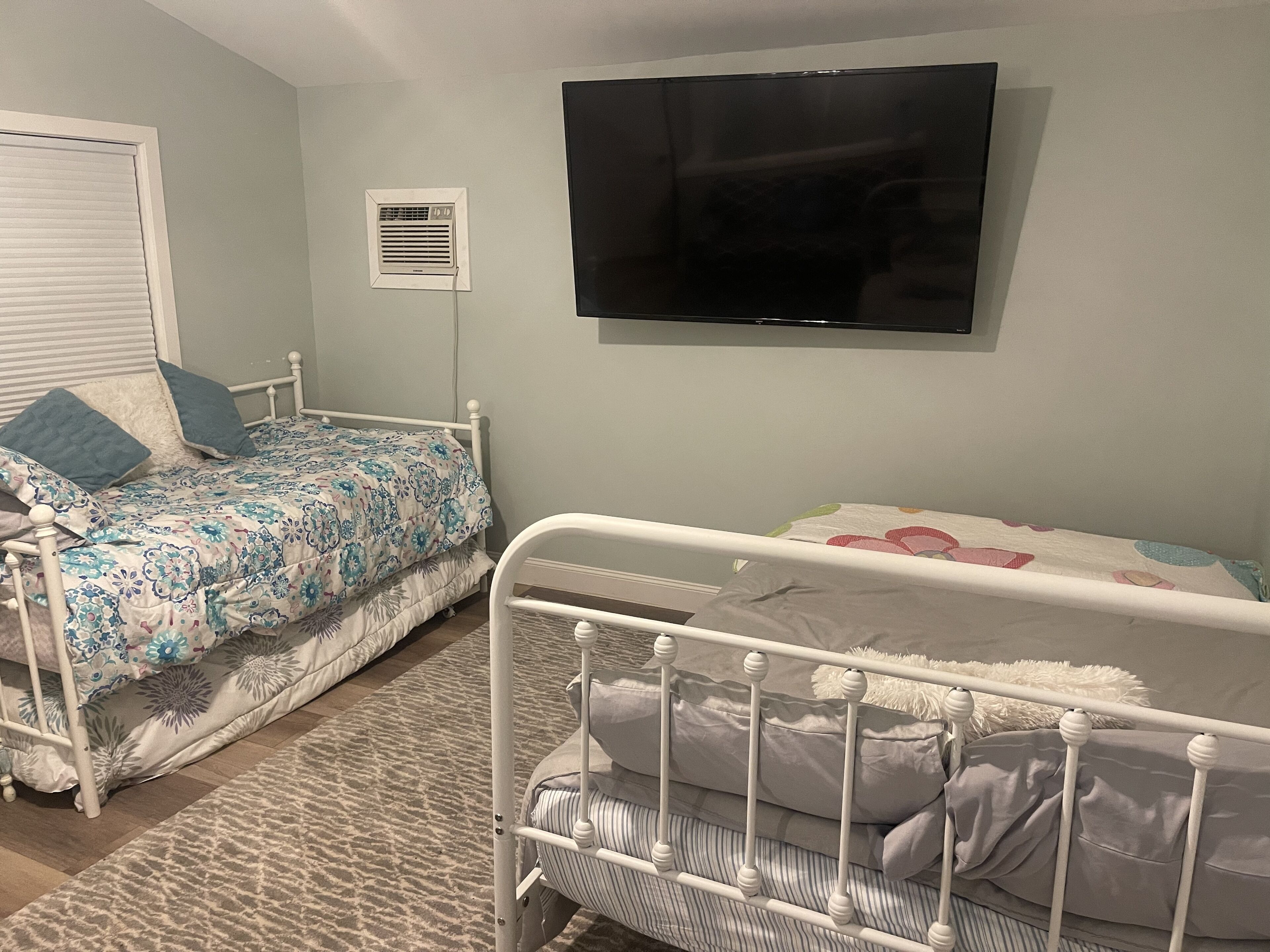 A bedroom with two beds, a TV mounted on the wall, and a window with blinds.