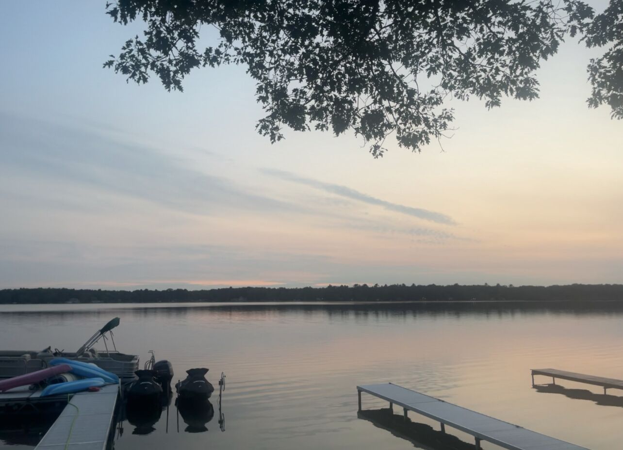 A calm lake with a dock and boats, surrounded by trees and a clear sky.