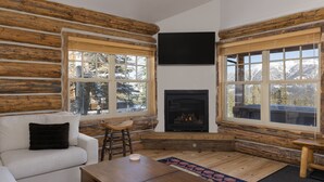 A log cabin interior with a fireplace, a sofa, a coffee table, and large windows offering a mountain view. - Derringer Haven | MTN Views, Hot Tub & Ski Access (Big Sky)