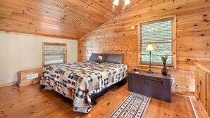 A cozy bedroom with a wooden ceiling, a bed with a checkered blanket, a nightstand, a lamp, and a window with blinds. - Almost Heaven Cabin - River Access! (Murphy)