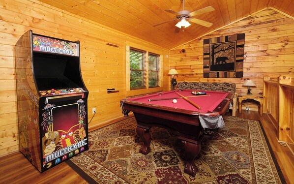 A wooden room with a pool table, a ceiling fan, and a video game machine.