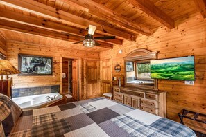 Ein Holzblock-Interieur mit Bett, Badezimmer mit Waschbecken und Spiegel, Deckenventilator und Fernseher an der Wand.