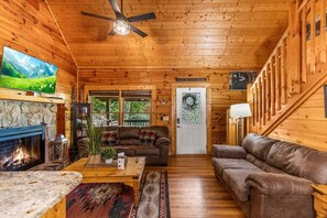 L’intérieur d’un chalet accueillant, avec une cheminée en pierre, des murs en bois, un ventilateur de plafond et un téléviseur accroché au mur gauche.