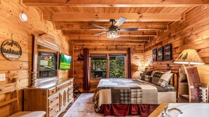 Interior de una cabaña con cama, televisor, ventana con vistas, ventilador de techo y piso de madera.