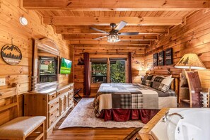 Ein Blockhaus-Interieur mit einem Bett, einem Fernseher, einem Fenster mit Aussicht, einem Deckenventilator und einem Holzboden.