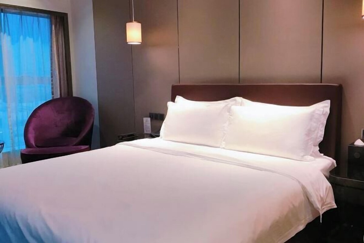 A hotel room with a large bed, a purple chair, and a nightstand with a lamp.