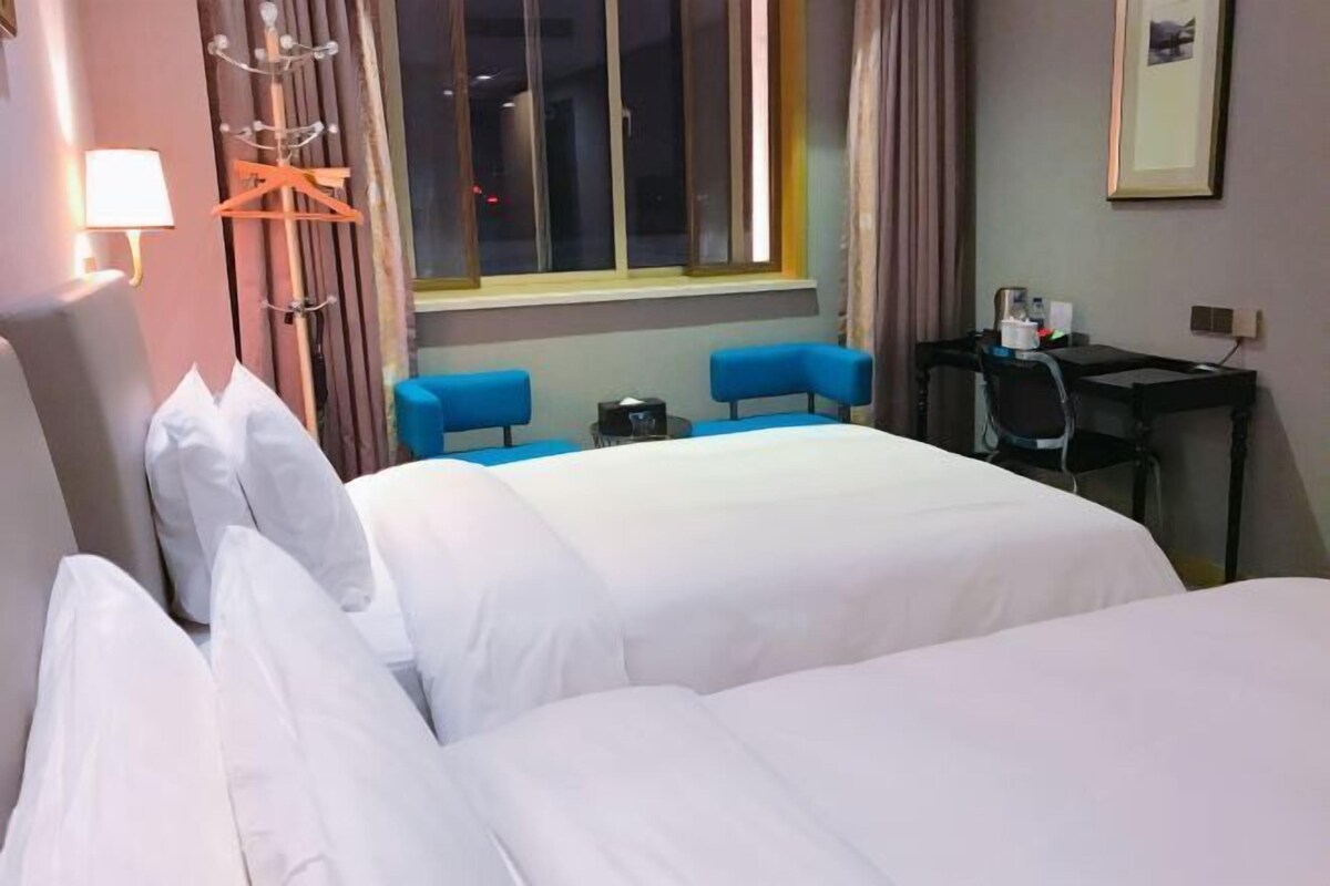 A hotel room with two beds, a desk, a chair, a lamp, a window with curtains, and a coat rack.