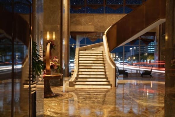 Eine moderne Hotelhalle mit Marmorboden, einer Treppe und Blick auf die beleuchtete Stadt bei Nacht.