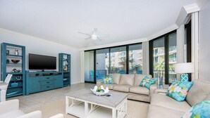 A modern living room with a flat-screen TV, teal cabinets, a beige sofa, and a large window offering a view of the city. - Stunning Views @ Phoenix on the Bay 1222 (Orange Beach)