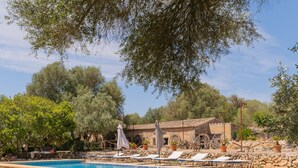 Un espace piscine doté de chaises longues et de parasols, entouré d’arbres et d’un bâtiment en pierre.