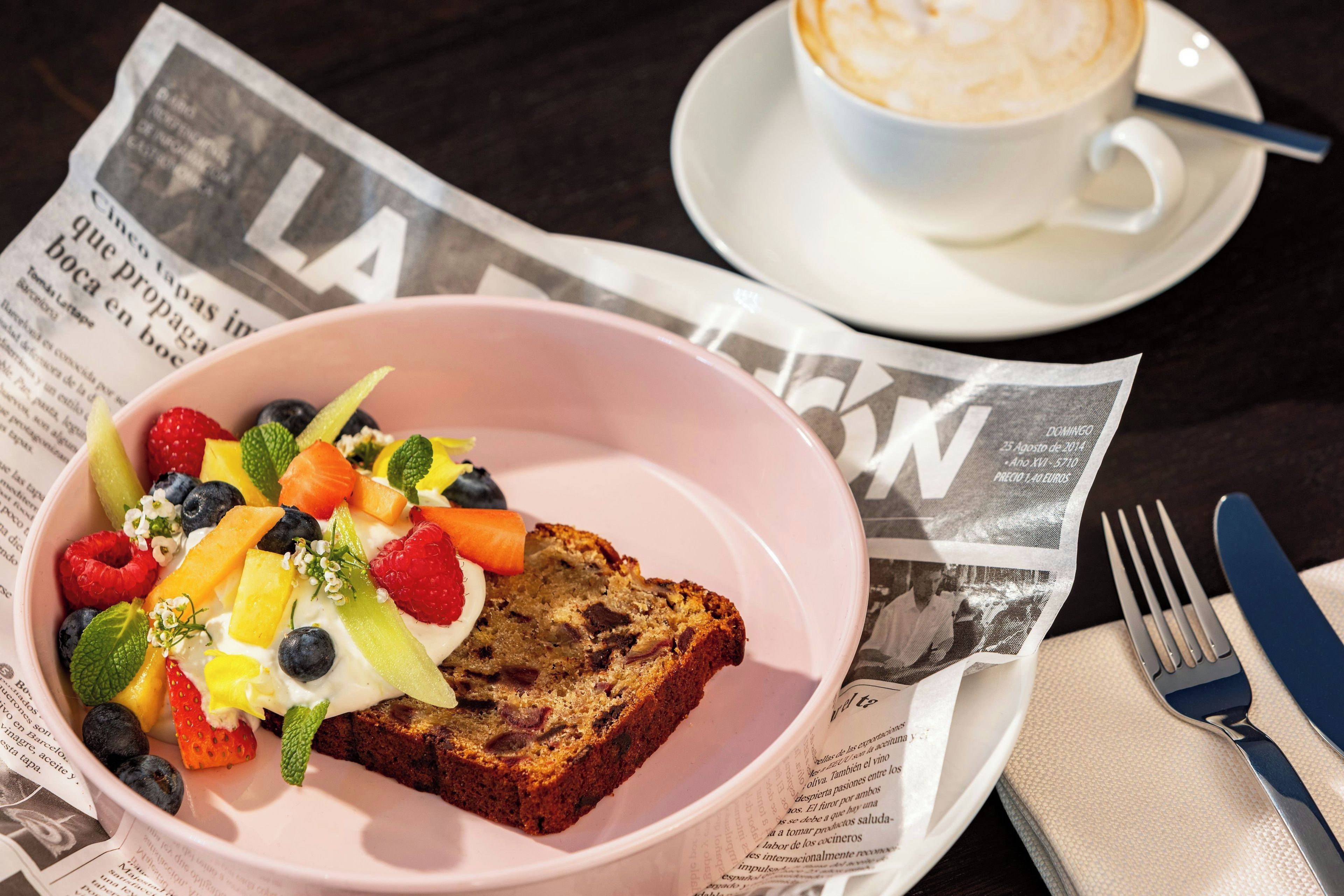 Un desayuno con un tazón de fruta, una rebanada de pan, una taza de café y un periódico sobre la mesa.