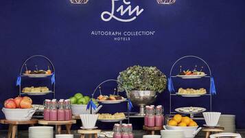 A buffet setup with various food items and beverages, including fruits, pastries, and drinks, displayed on tiered stands and tables against a dark blue wall with a logo and text.
