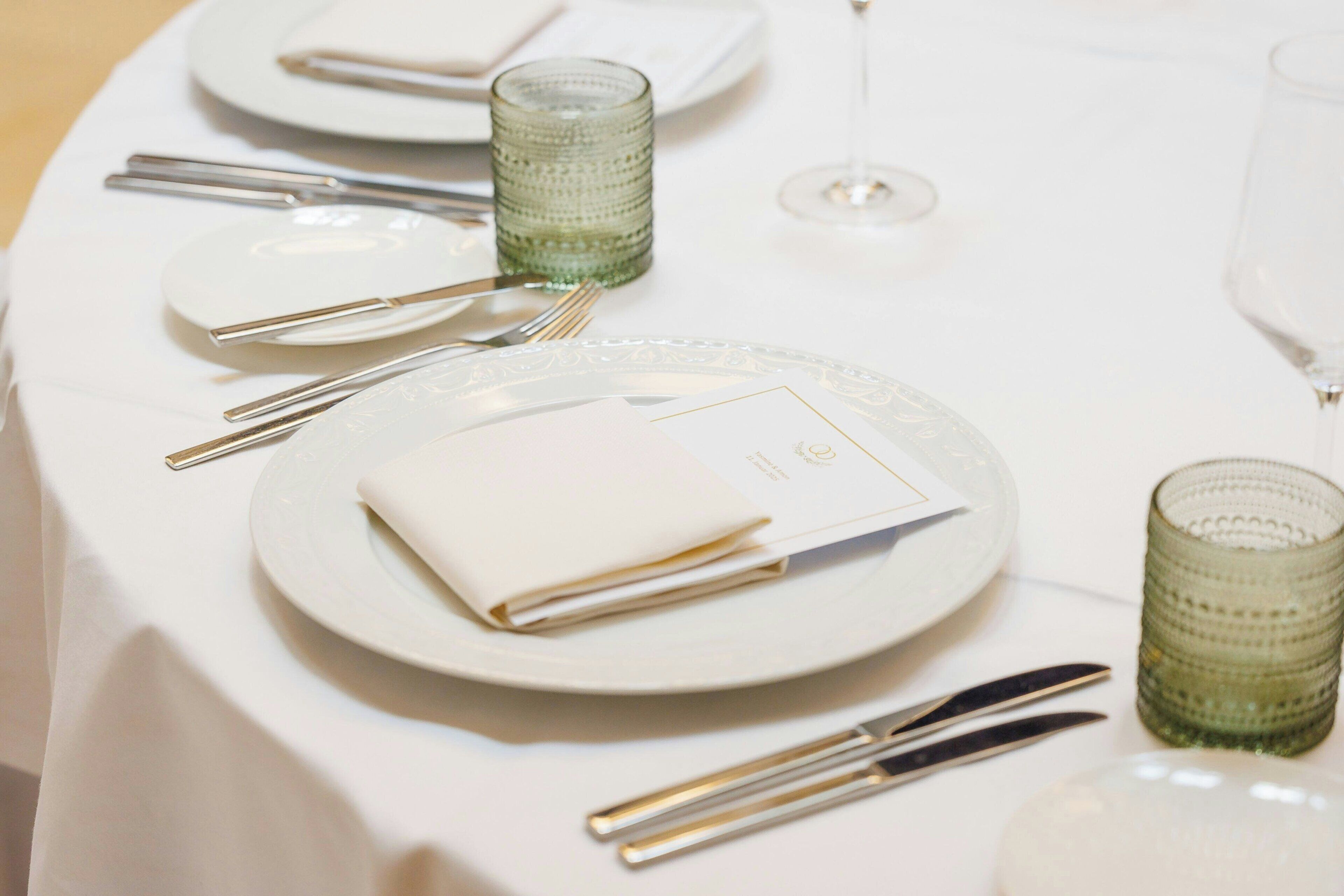 Una mesa servida con platos blancos, cubiertos y vasos de vidrio verde.