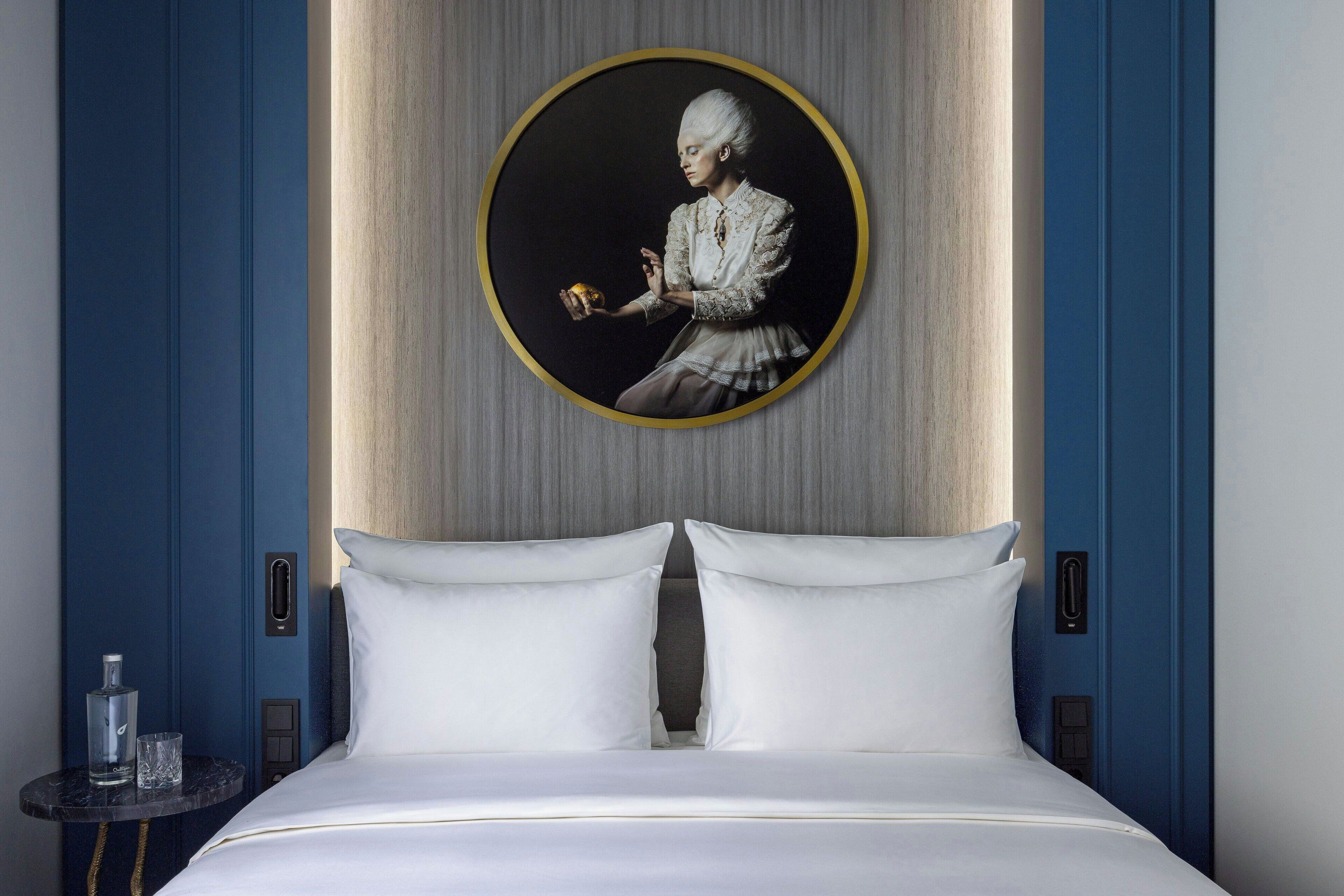 A hotel room with a large bed, two white pillows, a round portrait on the wall, and a small table with a bottle and glass.
