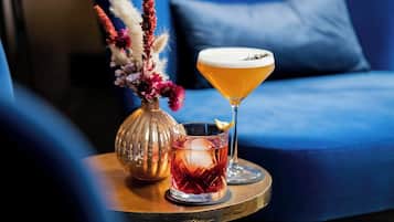A cocktail glass with a foamy head, a glass with a red liquid and ice cubes, and a vase with dried flowers on a table.
