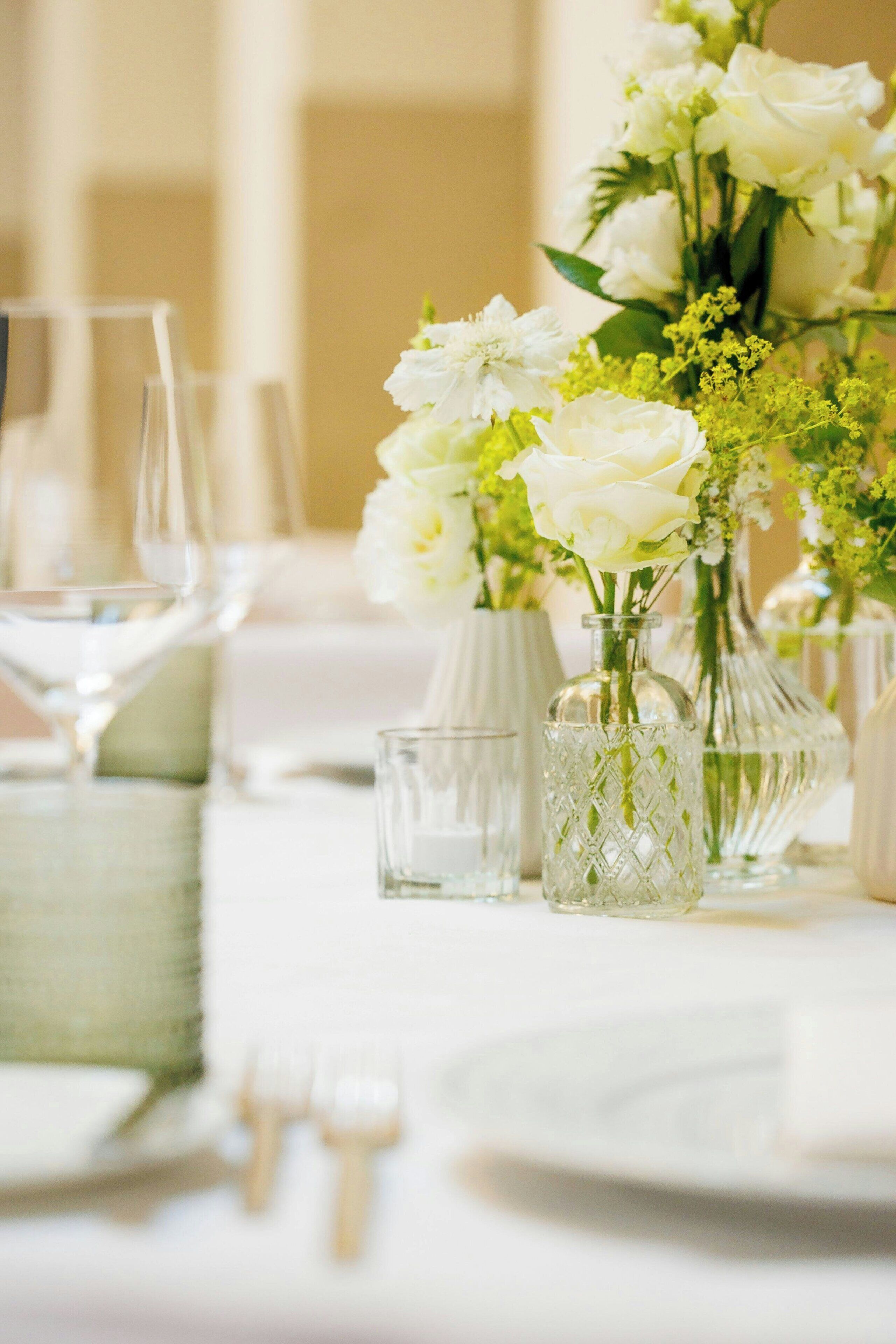 Een gedekte tafel met witte tafellinnen, heldere glazen vazen met witte bloemen en wijnglazen.