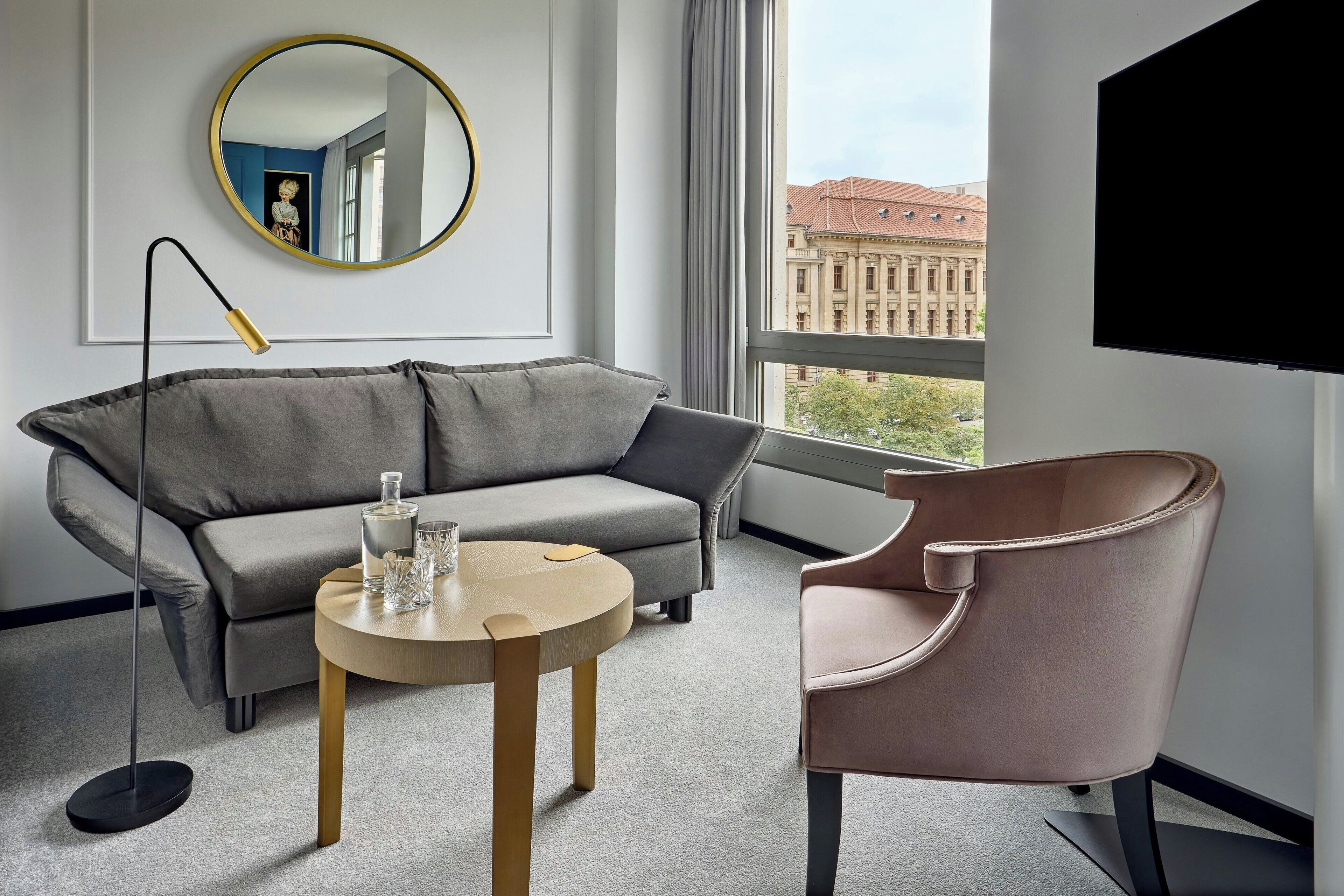 Une chambre d’hôtel moderne avec un canapé gris, une table basse ronde, un fauteuil rose, une lampe sur pied et une grande fenêtre donnant sur la ville.