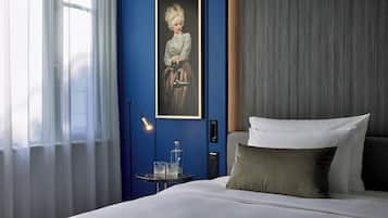 A hotel room with a bed, a bedside table with glasses and a bottle, a framed portrait on the wall, and a window with sheer curtains.