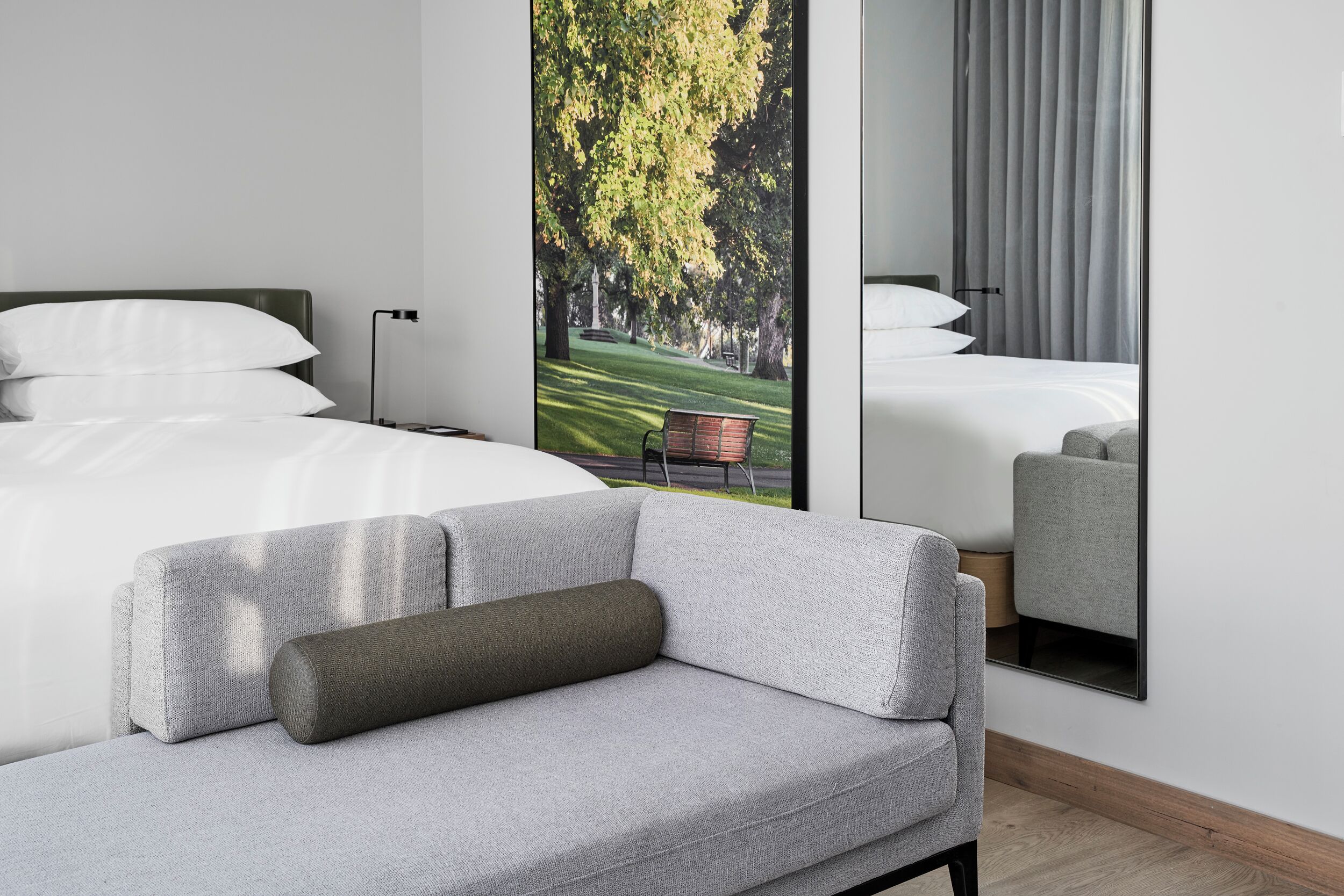 Ein modernes Hotelzimmer mit einem großen Bett, einem grauen Sofa, einem Spiegel und Blick auf einen Park durch ein Fenster.