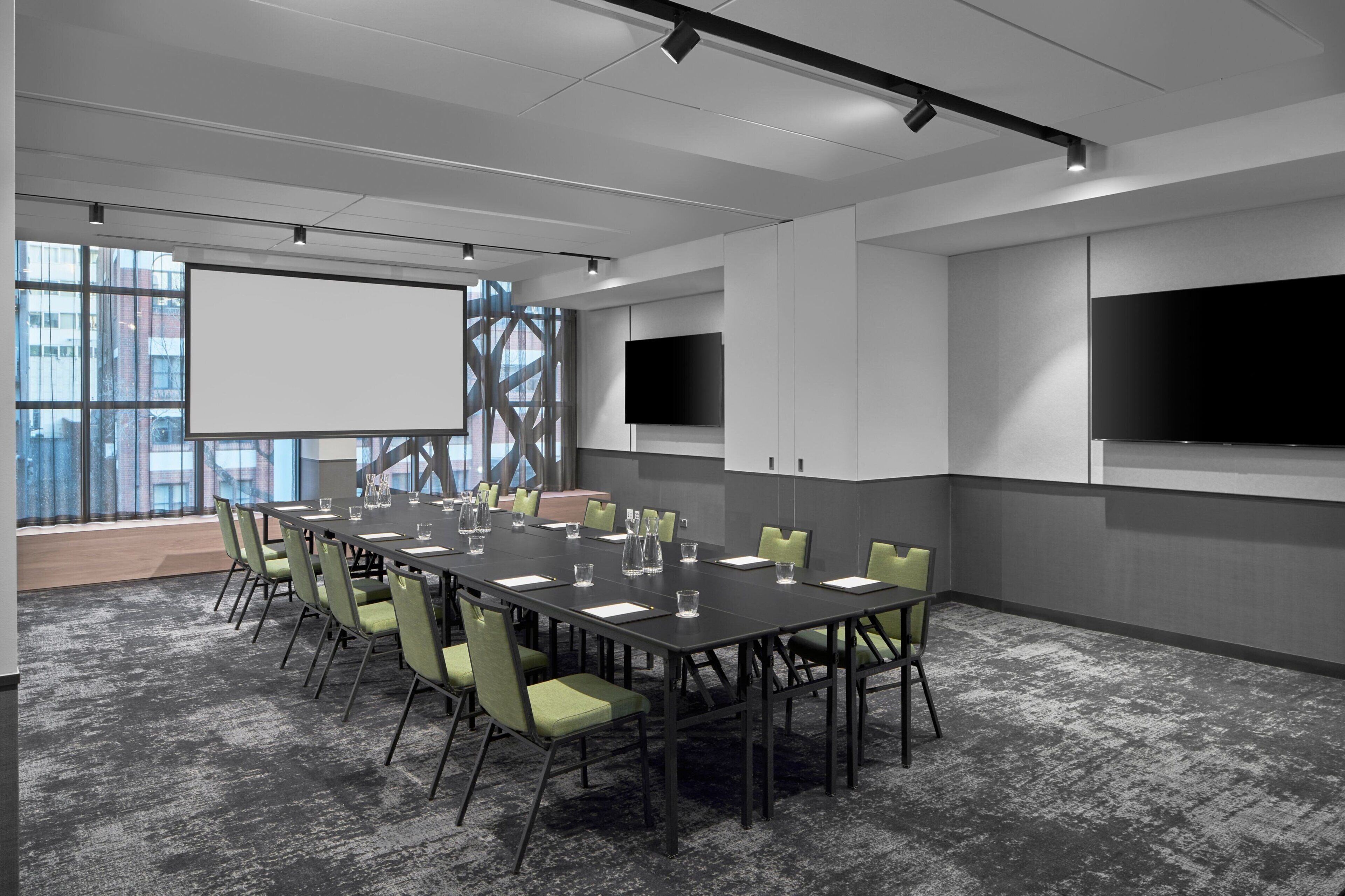 A conference room with a long table, green chairs, a projector screen, and two flat-screen TVs.