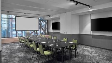 A conference room with a long table, green chairs, a projector screen, and two flat-screen TVs.