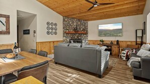 Un vaste espace de vie doté d’une cheminée en pierre, d’une table à manger avec des chaises, d’un canapé gris et d’un ventilateur de plafond.