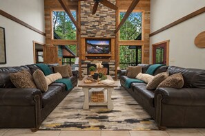 A spacious living room with two leather sofas, a stone fireplace, and a large window overlooking greenery. - Perfect for Family Gatherings, 4 King Suites, Heated Pool & Fire Pit (Broken Bow)
