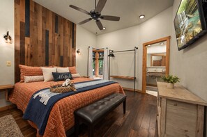 A bedroom with a bed, a bench, a ceiling fan, a wall-mounted TV, and a wooden floor. - Perfect for Family Gatherings, 4 King Suites, Heated Pool & Fire Pit (Broken Bow)