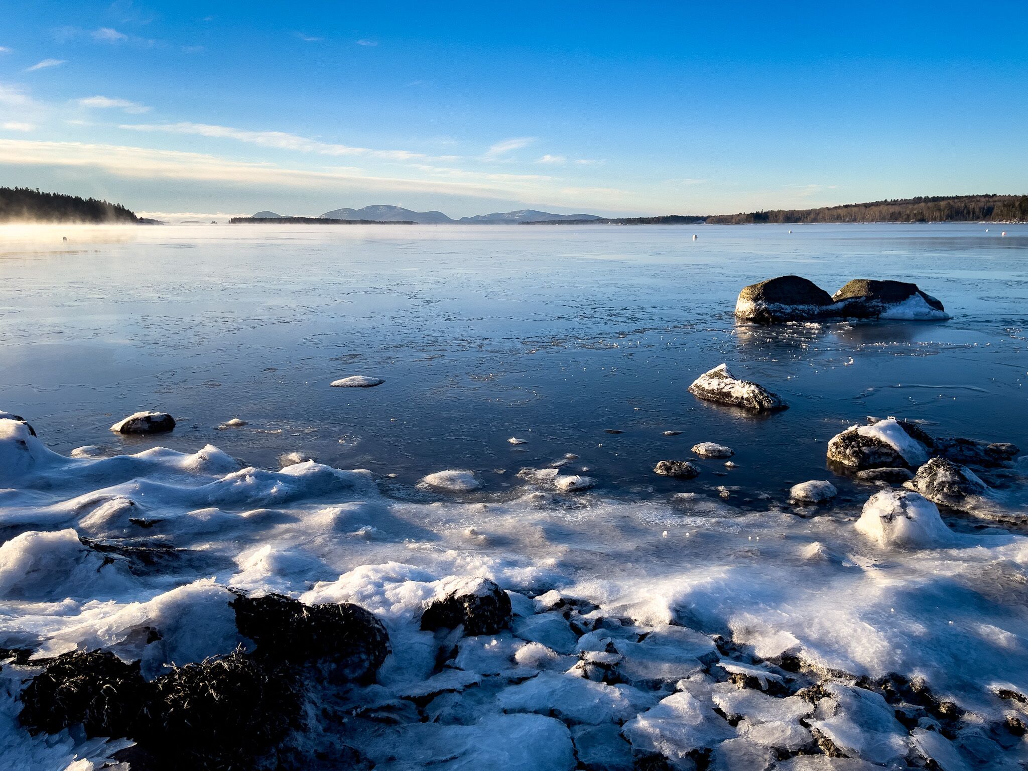 A frozen lake with ice formations and rocks, surrounded by trees and distant mountains.