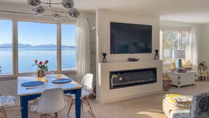 A living room with a fireplace, a dining table with chairs, a TV mounted on the wall, and a view of the water outside. - Lucky Stone Retreat - Private Beach & Acadia View (Sullivan)