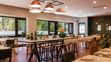 Un restaurant amb taules altes i tamborets de bar, finestres grans amb vista a vegetació, il·luminació penjant i una paret d'accent verd.
