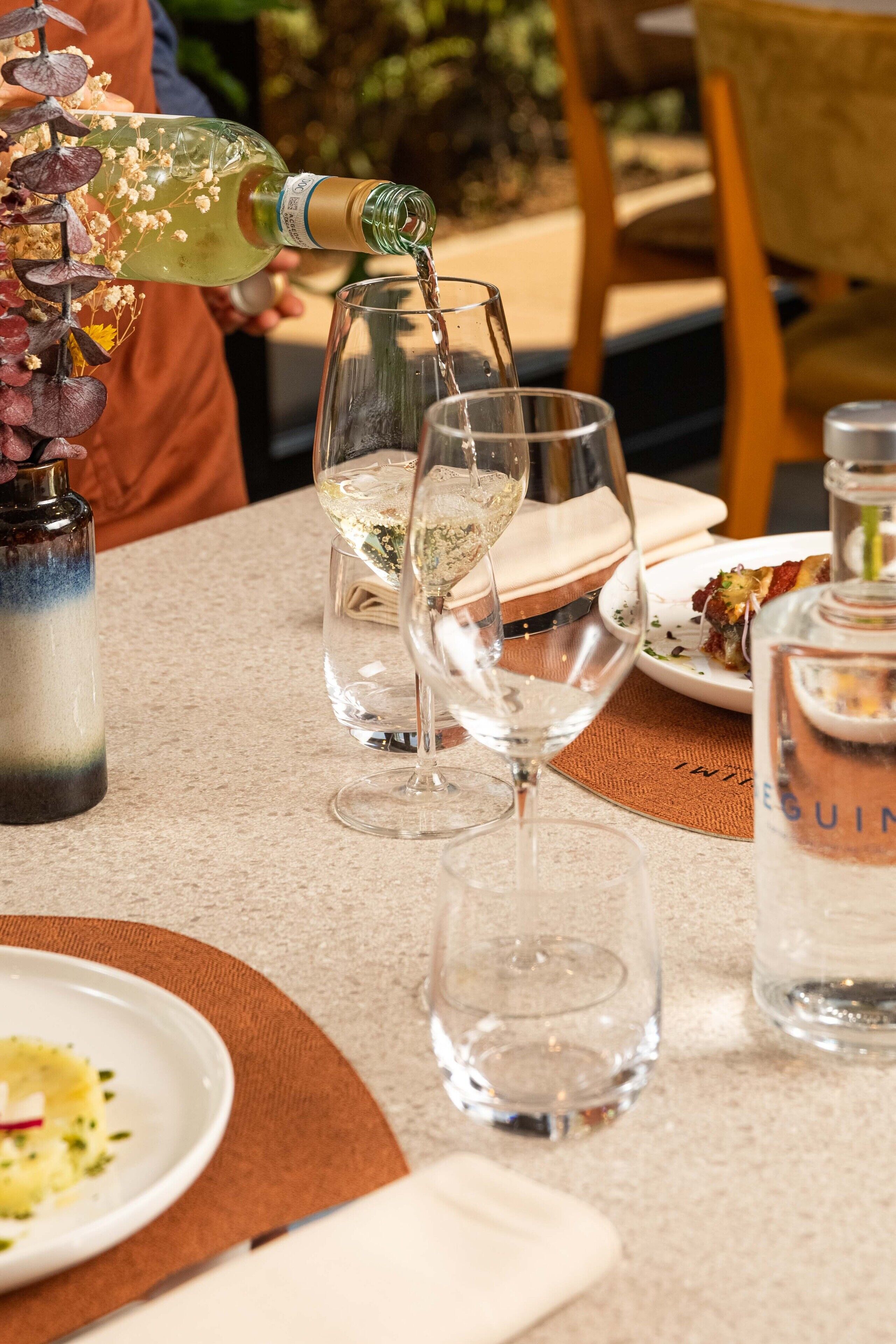 Una mesa con copas de vino, una botella de vino y un florero con flores secas.